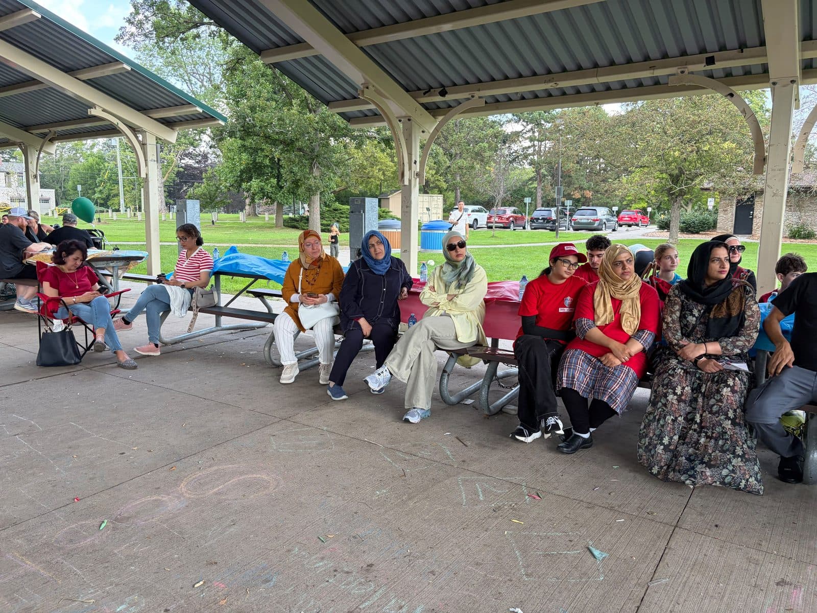 Families seated along picnic benches under a lakeshore park pavilion, some in red shirts