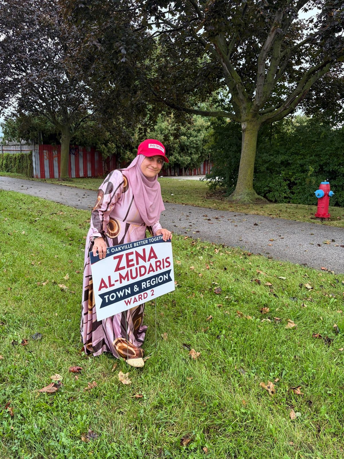 A woman in a red campaign cap kneels on a grassy boulevard to place a Zena Al-Mudaris, Town and Region, Ward 2 lawn sign